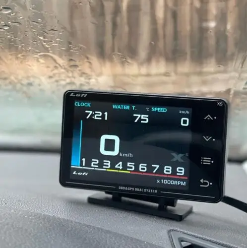 A Lufi Xs gauge on a car dashboard behind a rain-covered windscreen, showing clock, water temperature and speed